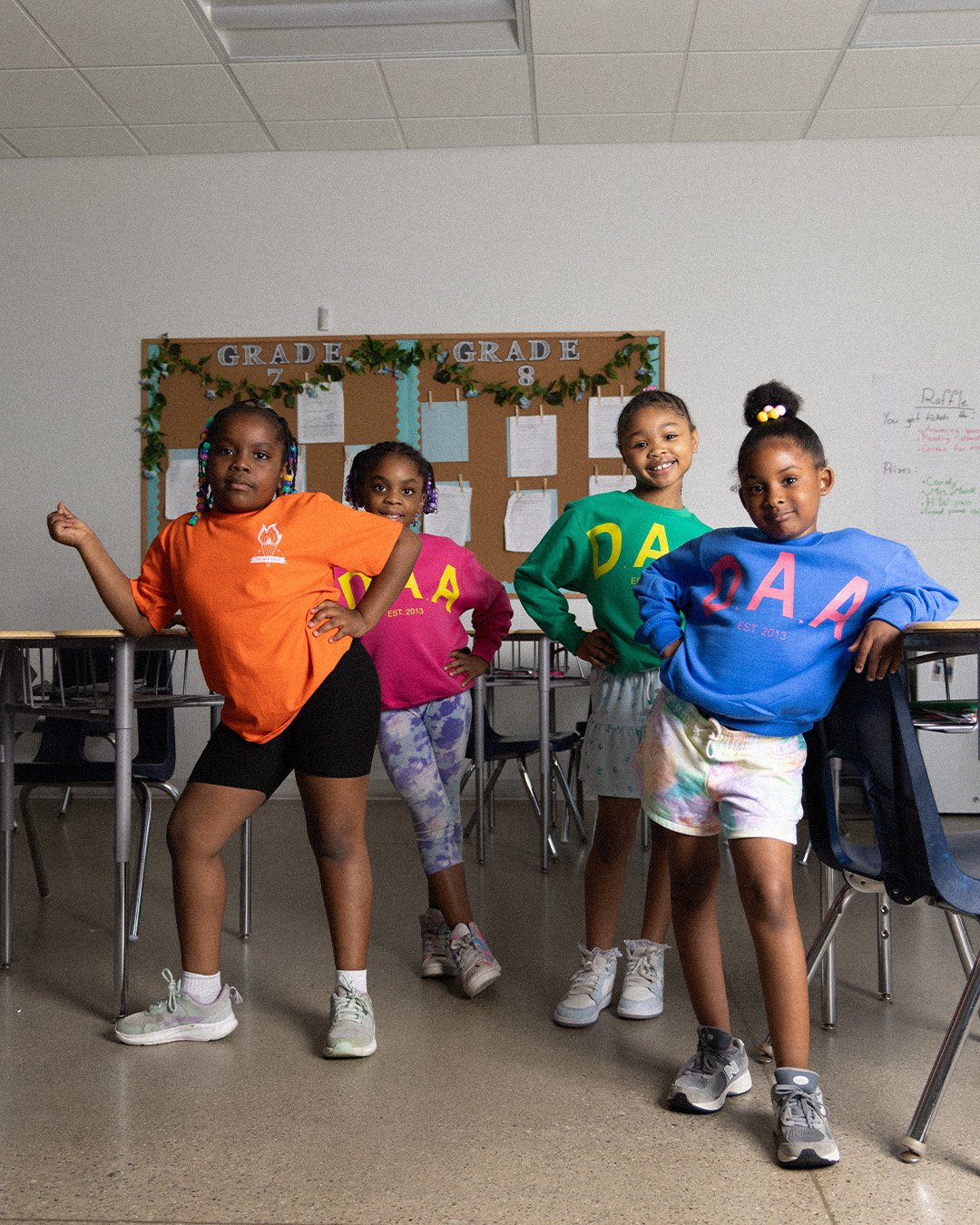 Four students posing in a classroom in the orange We Are Crew tee and pink, green and blue D.A.A. crewnecks