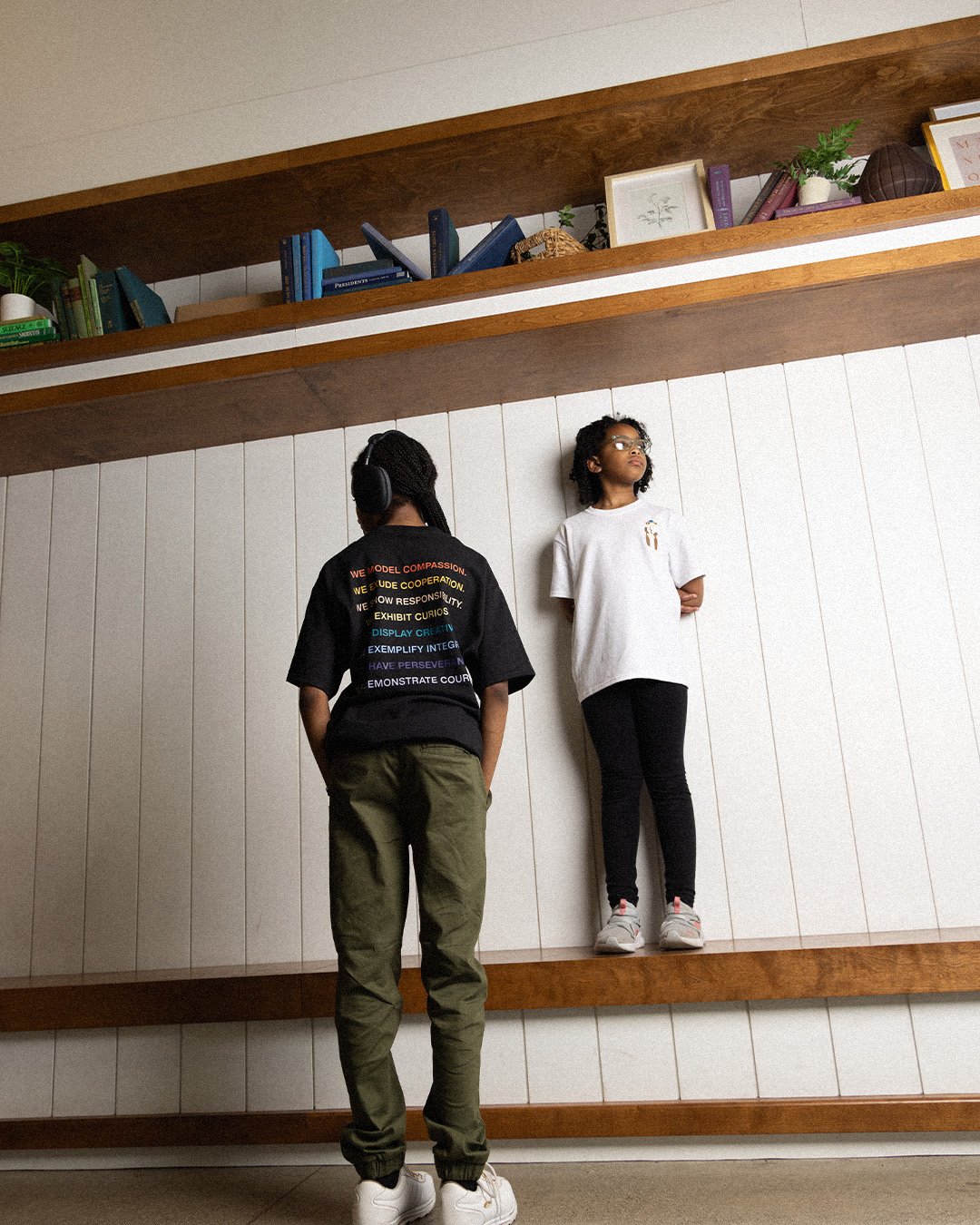 Two students by the library shelves, one wearing a black tee printed with the school values in rainbow colors